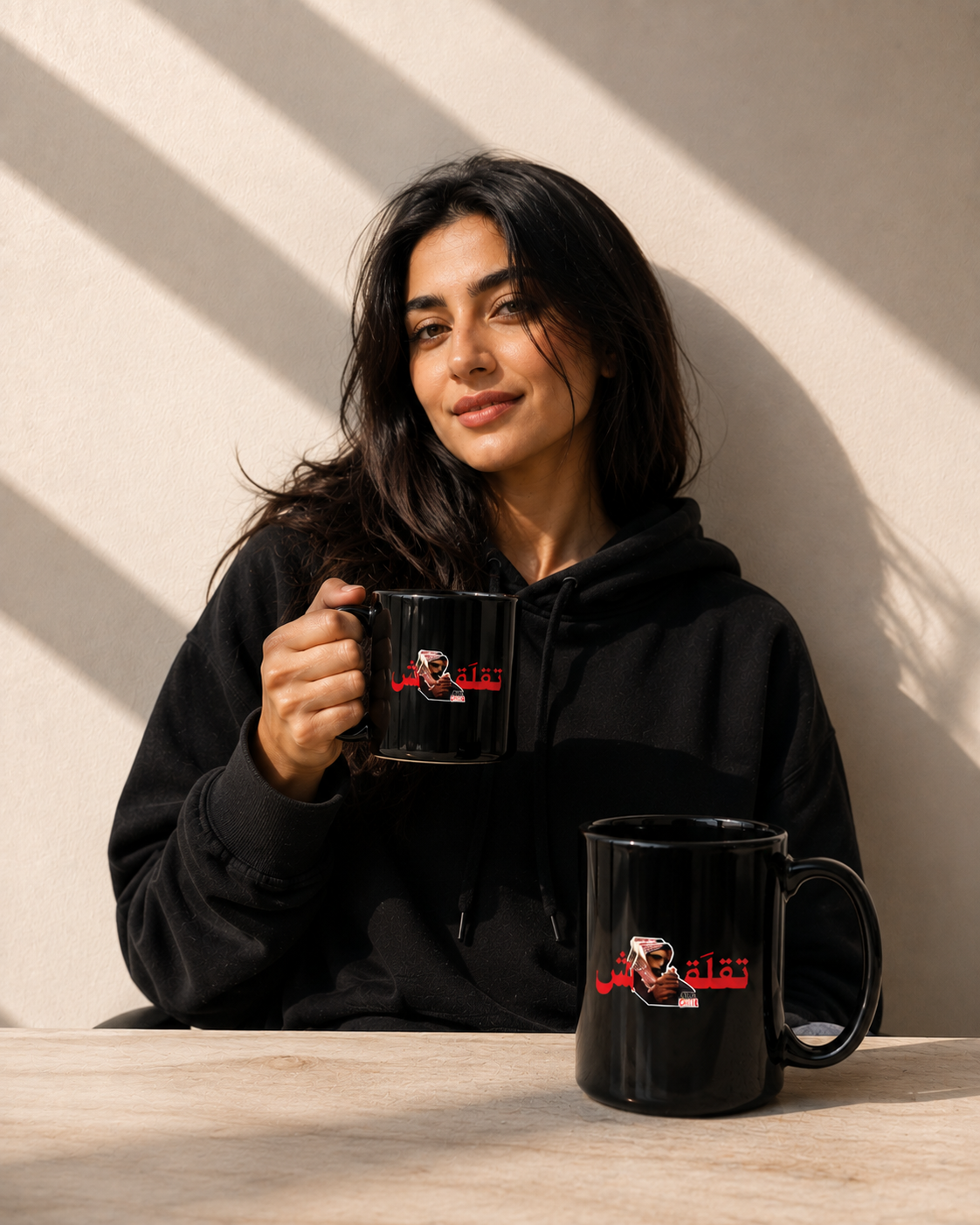 A woman in a black hoodie holds a black ceramic mug with a printed graphic, while another matching mug sits on a table in front of her against a sunlit beige wall.