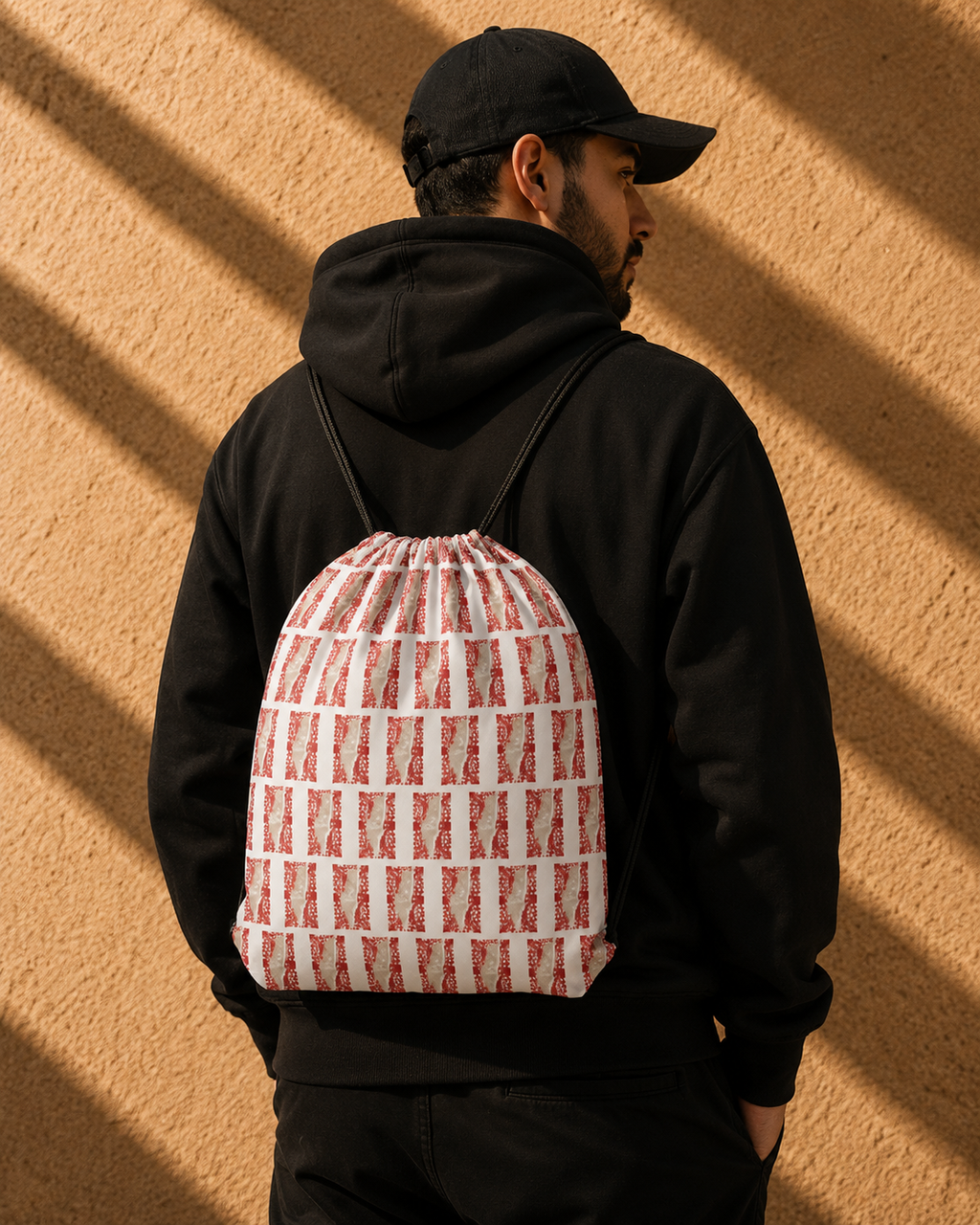 A man wearing a black hoodie and cap, viewed from behind, carrying a drawstring backpack with a red and white geometric pattern against a textured wall with diagonal shadows.