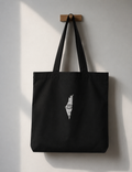 A black canvas tote bag with long shoulder straps hangs from a small wooden peg on a light gray wall. A white graphic of the map of Palestine is printed on the front of the bag.