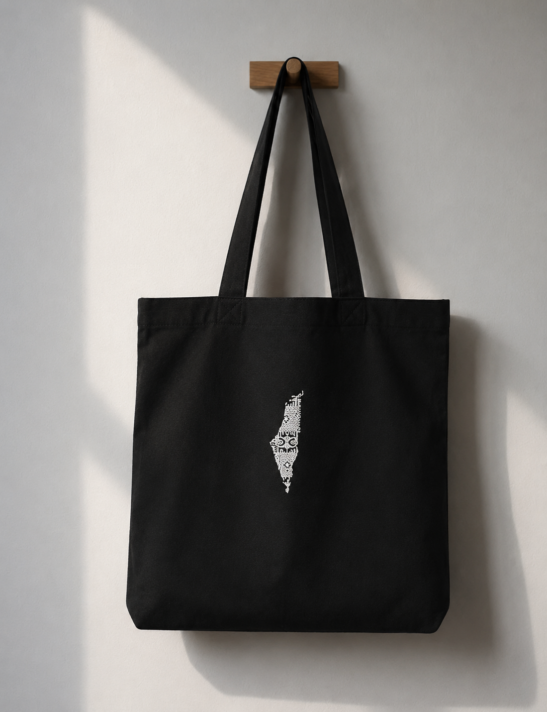 A black canvas tote bag with long shoulder straps hangs from a small wooden peg on a light gray wall. A white graphic of the map of Palestine is printed on the front of the bag.