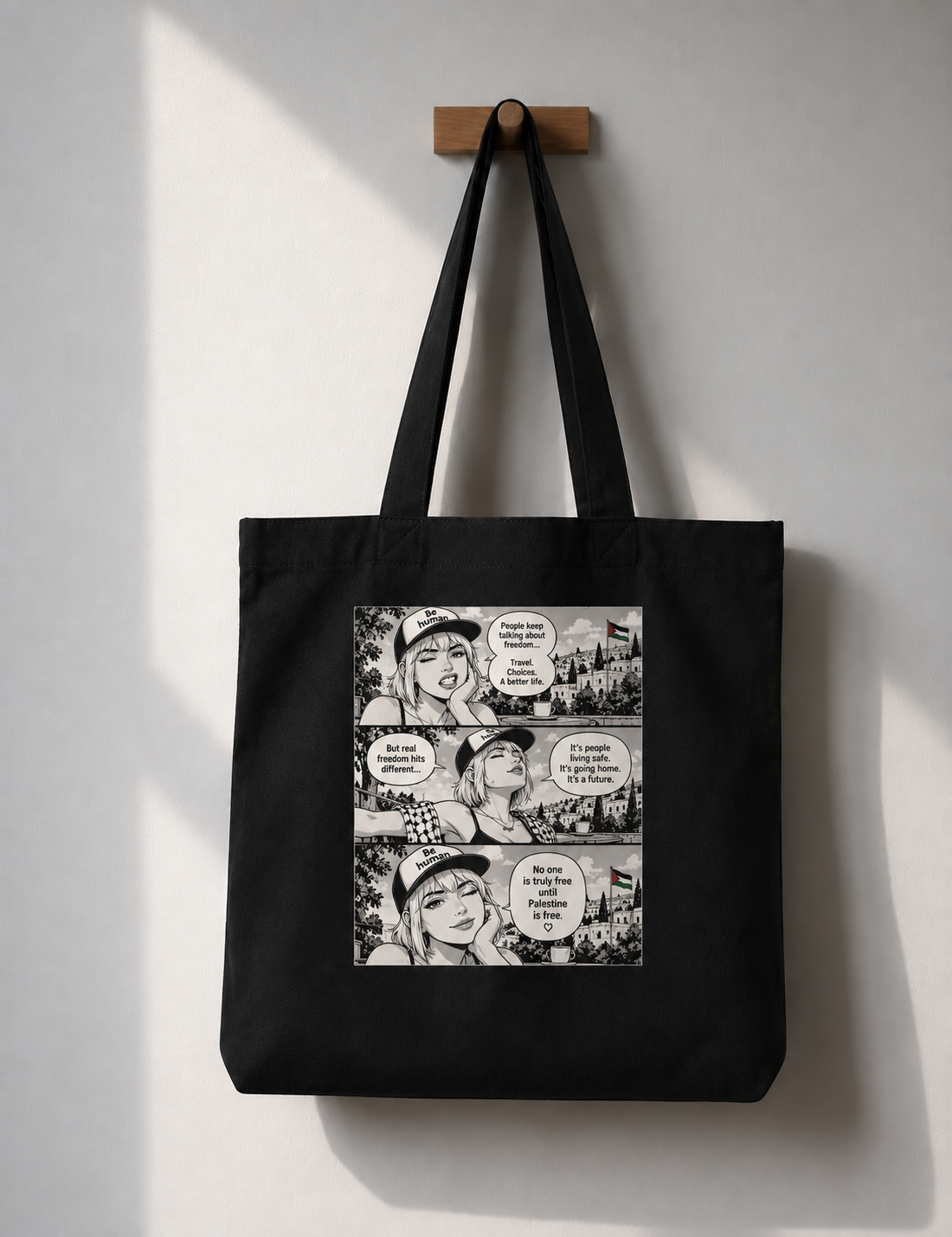 A black canvas tote bag hanging from a wooden peg on a light gray wall. The bag features a large rectangular graphic print on the front with a black-and-white manga comic-style illustration and speech bubbles.