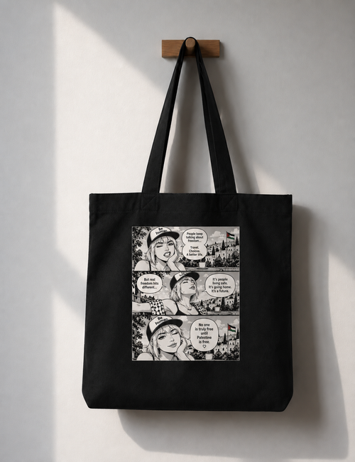 A black canvas tote bag hanging from a wooden peg on a light gray wall. The bag features a large rectangular graphic print on the front with a black-and-white manga comic-style illustration and speech bubbles.