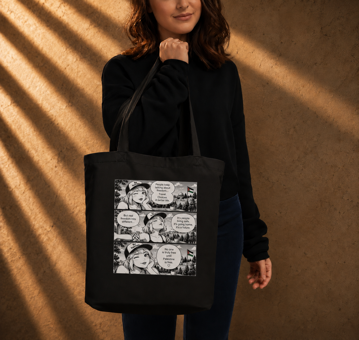 A woman wearing a black sweatshirt and dark pants carries a black canvas tote bag with a black-and-white comic strip graphic on the front, standing against a warm brown textured wall with dramatic diagonal shadows.