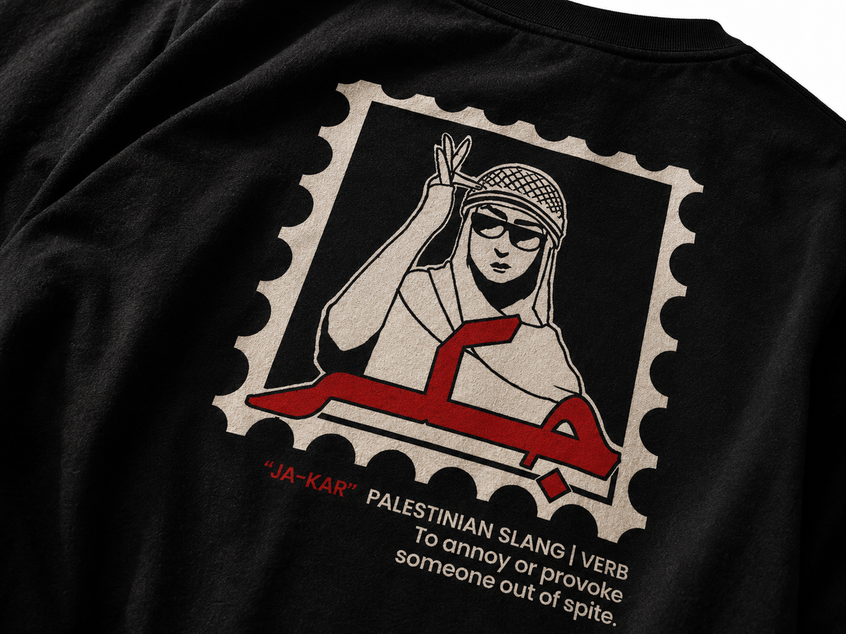 Close-up of a black T-shirt featuring a large graphic print on the back. The print resembles a postage stamp with a stylized illustration of a person in a headscarf and sunglasses making a peace sign, with a red Arabic script element and text reading "'JA-KAR' PALESTINIAN SLANG | VERB To annoy or provoke someone out of spite.