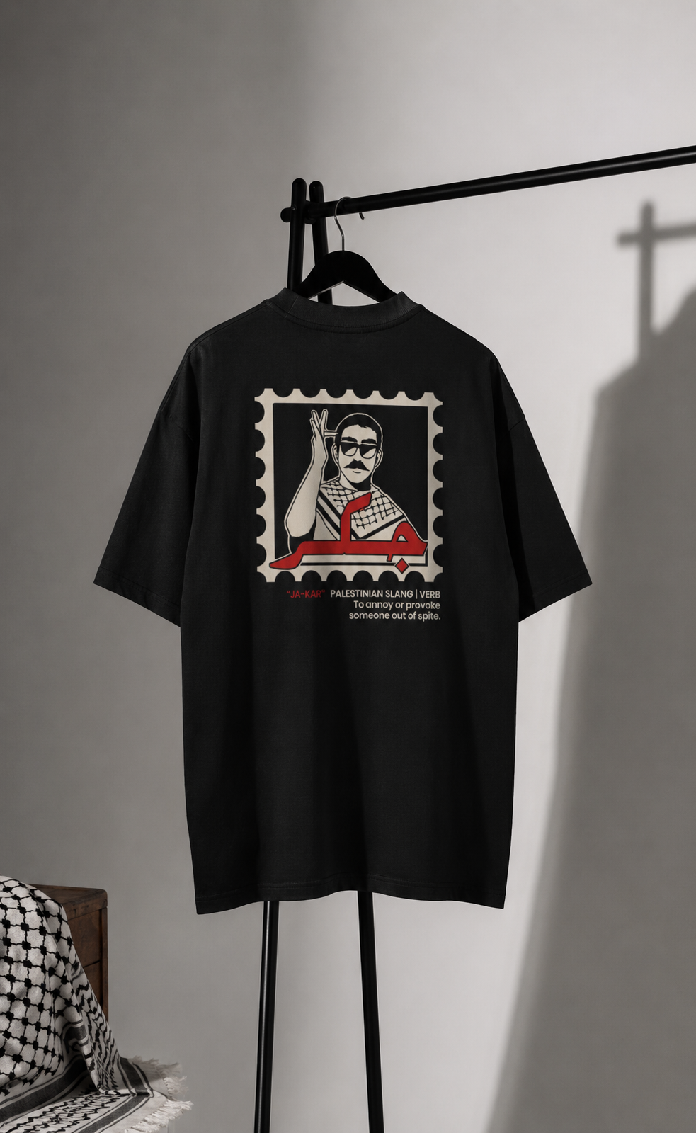 A close-up of a black t-shirt featuring a graphic print of a postage stamp. The stamp contains a stylized illustration of a man raising his hand, with red Arabic calligraphy and the text '*JA-KAR' 'PALESTINIAN SLANG | VERB To annoy or provoke someone out of spite.