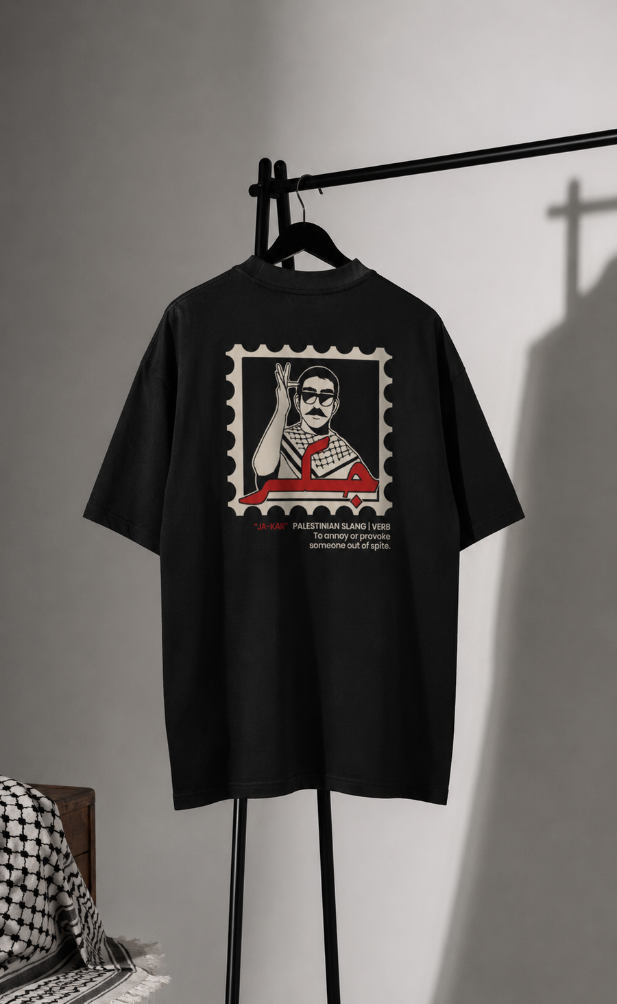 A close-up of a black t-shirt featuring a graphic print of a postage stamp. The stamp contains a stylized illustration of a man raising his hand, with red Arabic calligraphy and the text '*JA-KAR' 'PALESTINIAN SLANG | VERB To annoy or provoke someone out of spite.