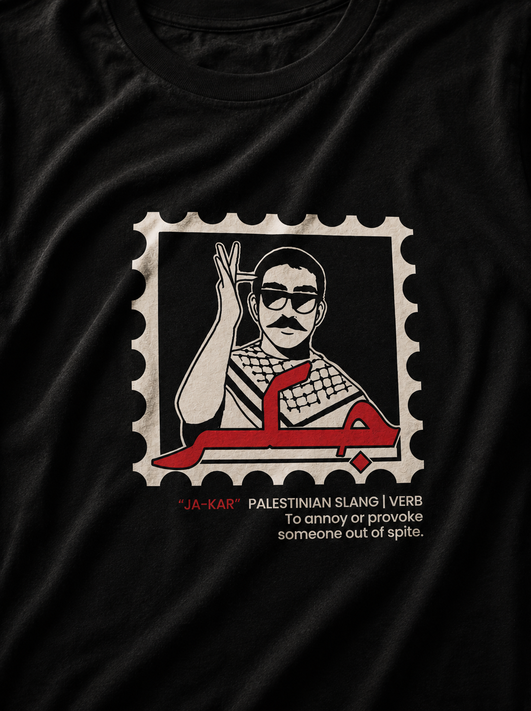 A close-up of a black t-shirt featuring a graphic print of a postage stamp. The stamp contains a stylized illustration of a man raising his hand, with red Arabic calligraphy and the text '*JA-KAR' 'PALESTINIAN SLANG | VERB To annoy or provoke someone out of spite.