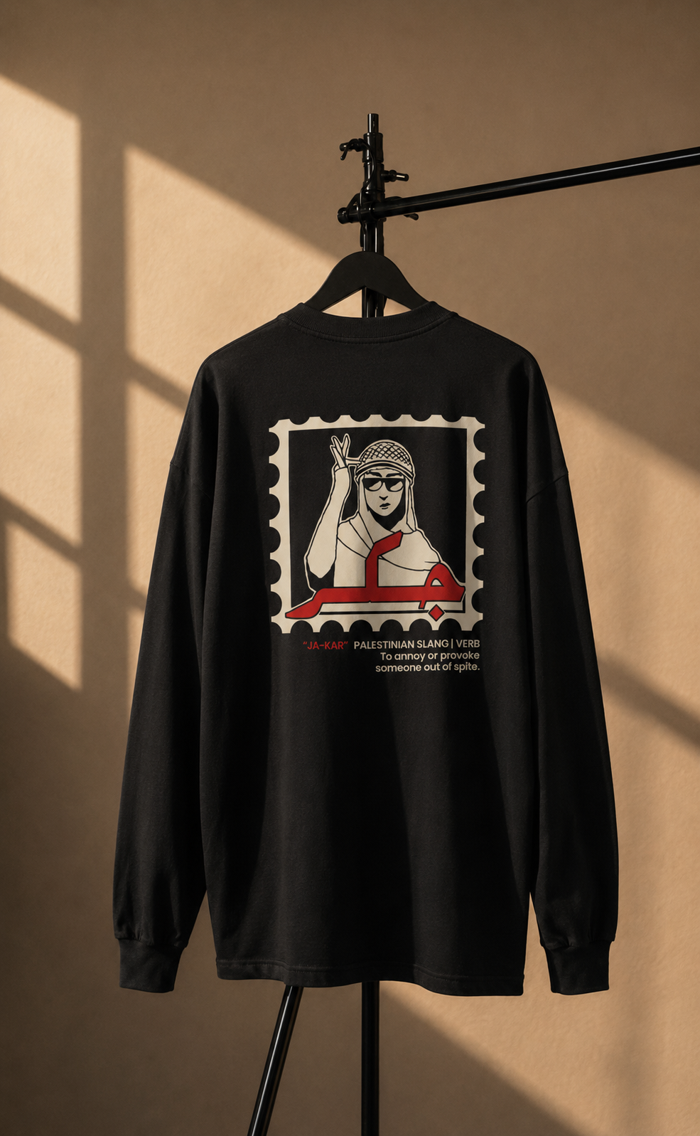 A black long-sleeve t-shirt hanging on a black hanger stand against a warm beige wall with dramatic window shadow lighting. The back of the shirt features a large graphic print styled like a postage stamp, showing a stylized illustration of a woman in a headscarf and a red vehicle, with text below.