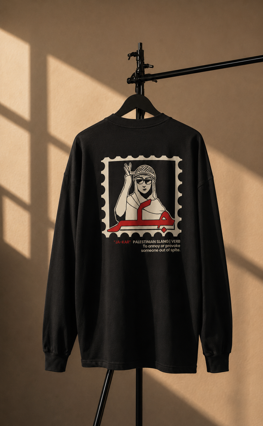 A black long-sleeve t-shirt hanging on a black hanger stand against a warm beige wall with dramatic window shadow lighting. The back of the shirt features a large graphic print styled like a postage stamp, showing a stylized illustration of a woman in a headscarf and a red vehicle, with text below.