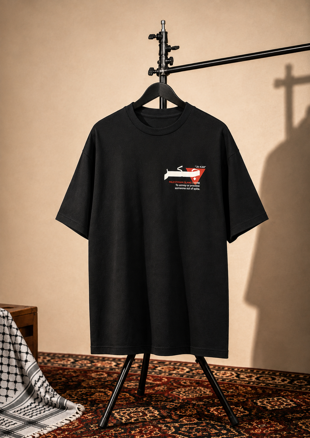 A black short-sleeve t-shirt hanging on a black hanger stand, featuring a small chest graphic of a red and white car with text. The setup is styled in a warm, rustic room with a beige wall, patterned rug, and a draped black-and-white patterned cloth on the left.