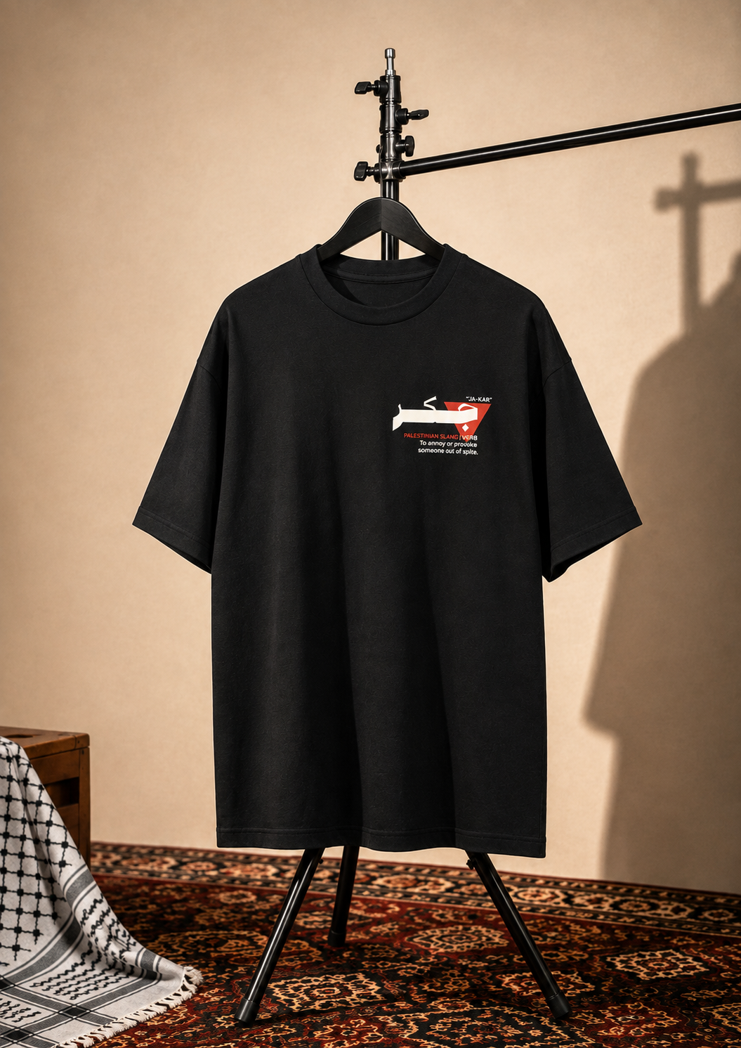 A black short-sleeve t-shirt hanging on a black hanger stand, featuring a small chest graphic of a red and white car with text. The setup is styled in a warm, rustic room with a beige wall, patterned rug, and a draped black-and-white patterned cloth on the left.