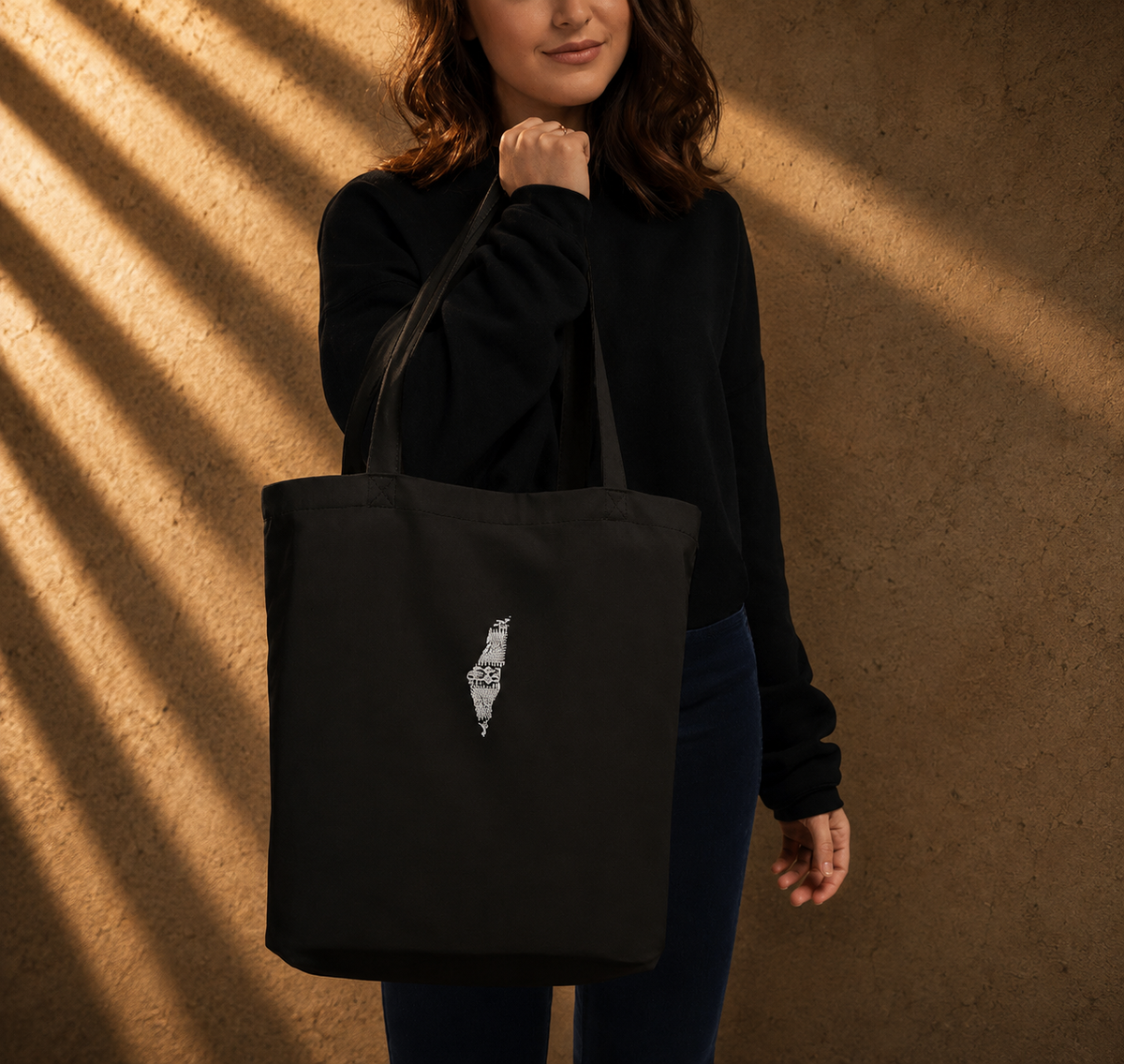 A woman wearing a black sweatshirt and dark pants carries a black canvas tote bag with a small white graphic of a hand holding a coffee cup on the front, set against a textured wall with warm sunlight shadows.