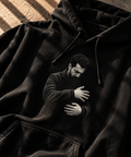 A black pullover hoodie laid flat on a textured surface, featuring a monochrome printed graphic of a man hugging himself in the center chest area.