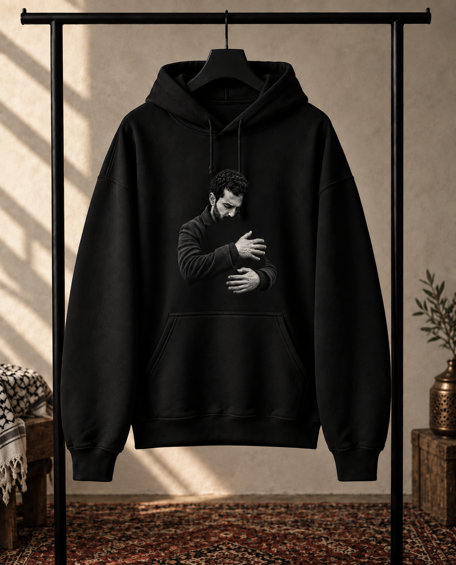A black pullover hoodie hanging on a black clothing rack, featuring a black-and-white graphic print of a man hugging another person on the front. The hoodie is set against a warm, rustic background with a textured wall, patterned rug, and small decorative plants.