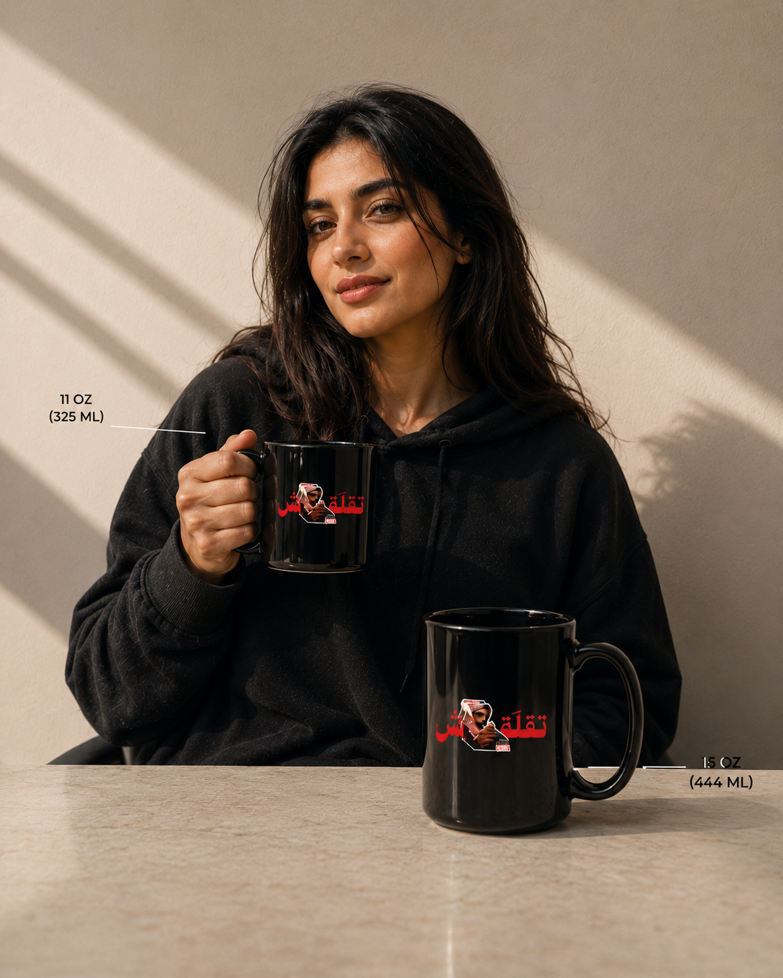 A woman in a black hoodie holds a black 11 oz mug while another black mug sits on a table in front of her. Both mugs feature a red Arabic text graphic and a portrait illustration.