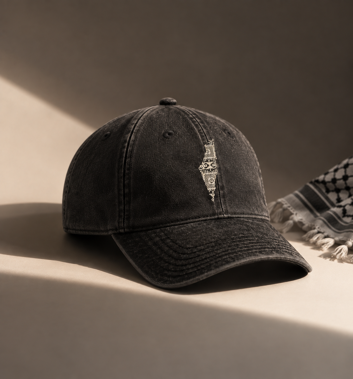 A washed black baseball cap with a distressed vertical logo on the front, styled on a warm beige surface with dramatic side lighting and a patterned scarf in the background.