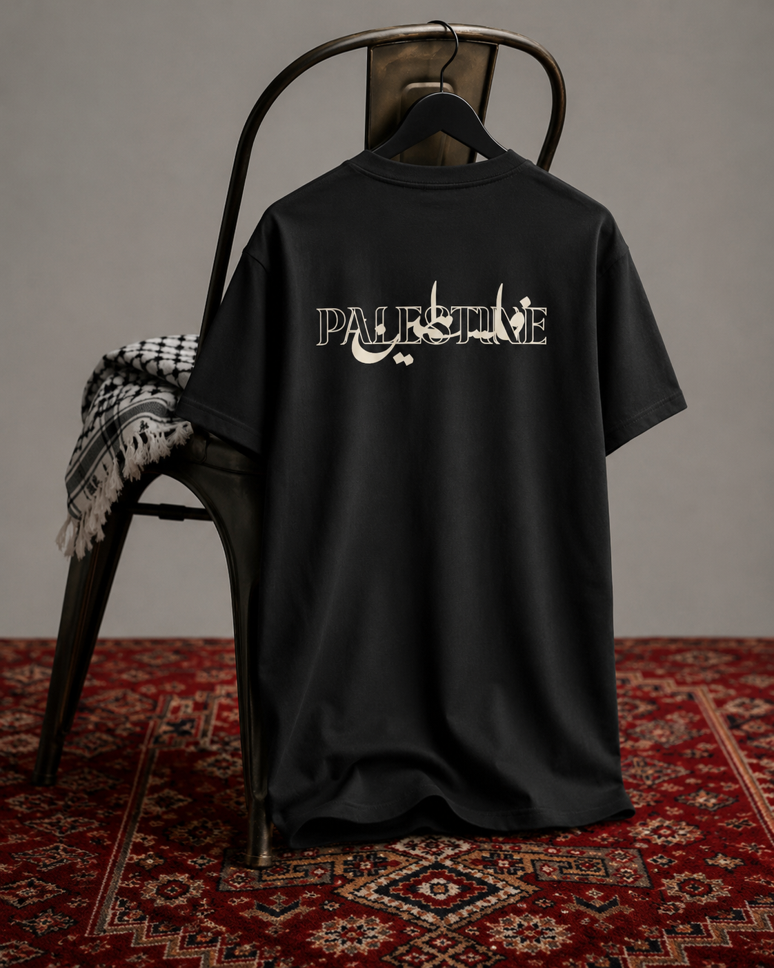 A black oversized T-shirt hanging on a chair, shown from the back with a large distressed white 'PALESTINE' graphic across the upper back. The shirt is styled in a studio setup with a dark metal chair, a patterned throw blanket, and a red patterned rug against a neutral gray wall.