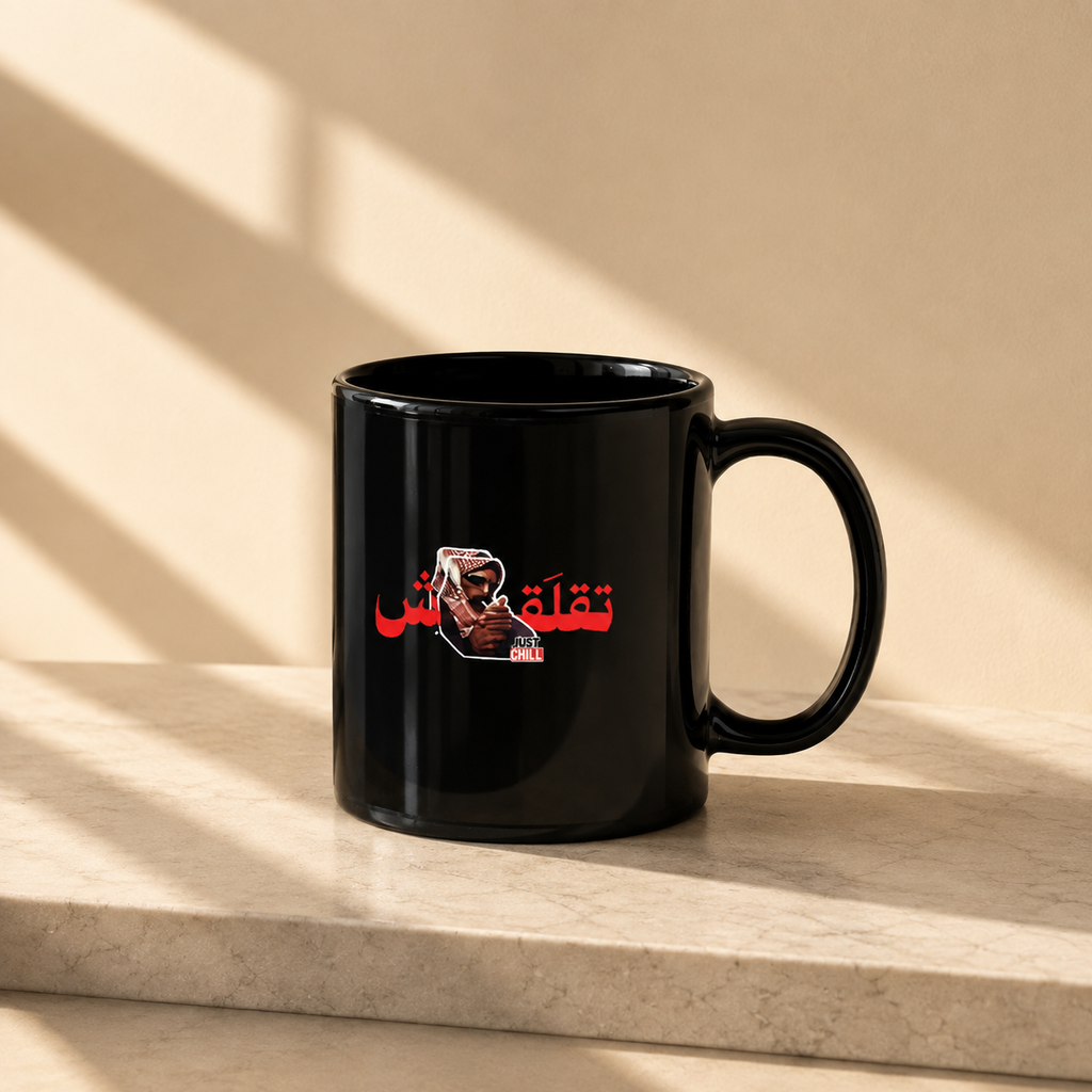 A glossy black ceramic mug with a red and white graphic print of Deadpool and Arabic text, sitting on a light beige stone ledge with diagonal window shadows cast across the background.