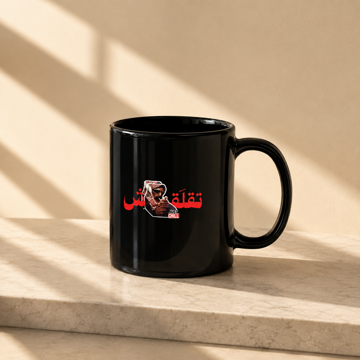 A glossy black ceramic mug with a red and white graphic print of Deadpool and Arabic text, sitting on a light beige stone ledge with diagonal window shadows cast across the background.