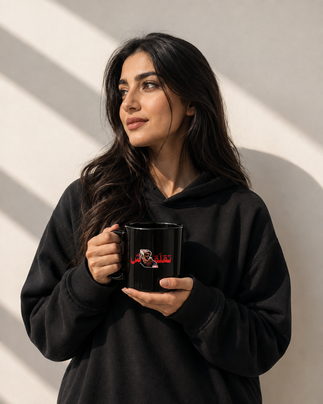 A woman with long dark hair wearing a black hoodie holds a black ceramic mug with a red and white graphic print. She is standing against a warm-toned wall with diagonal shadows.