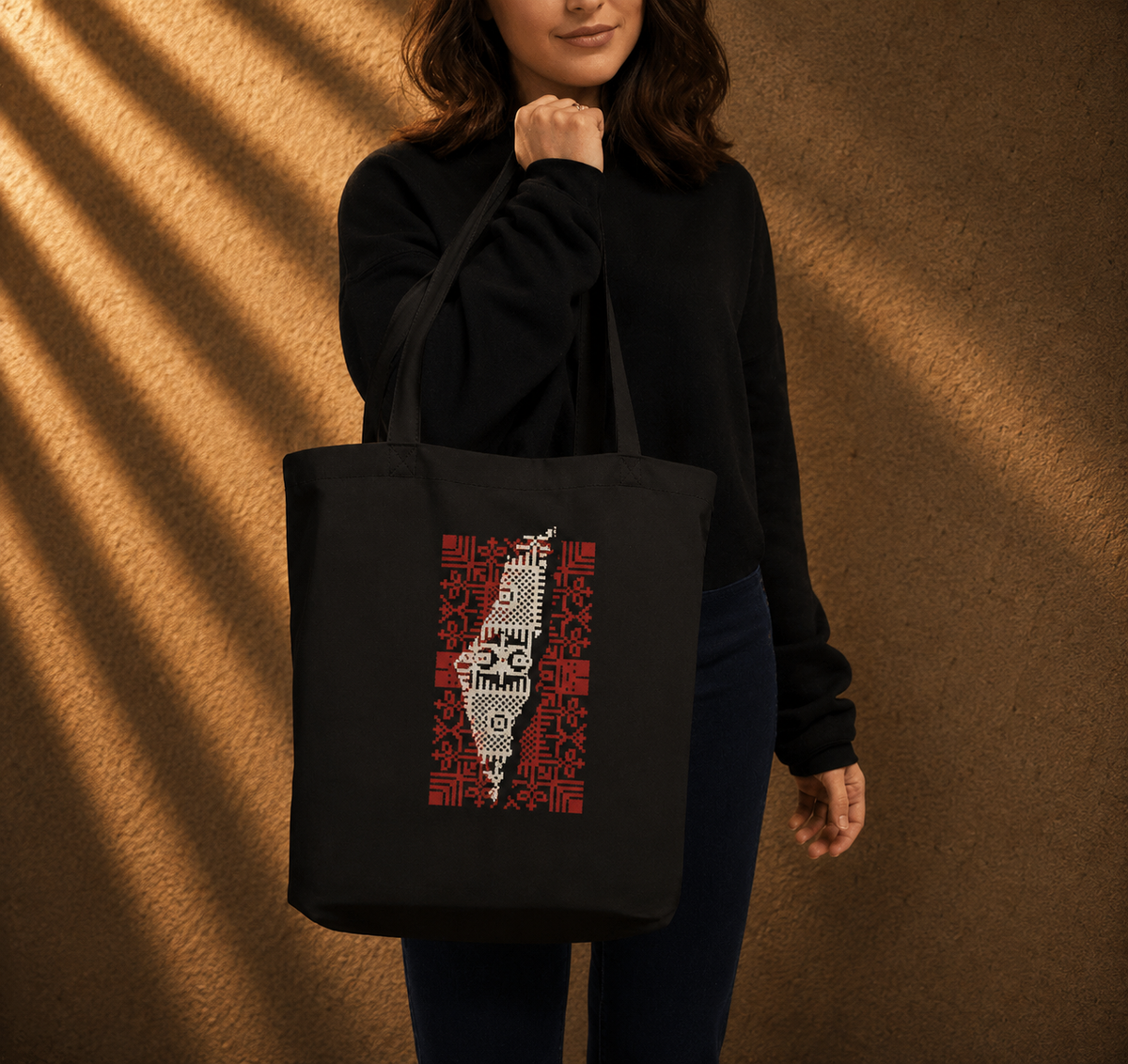 A woman wearing a black sweatshirt and dark jeans carries a black canvas tote bag with a red and white graphic print of the map of Palestine filled with text, set against a textured brown wall with dramatic diagonal shadows.