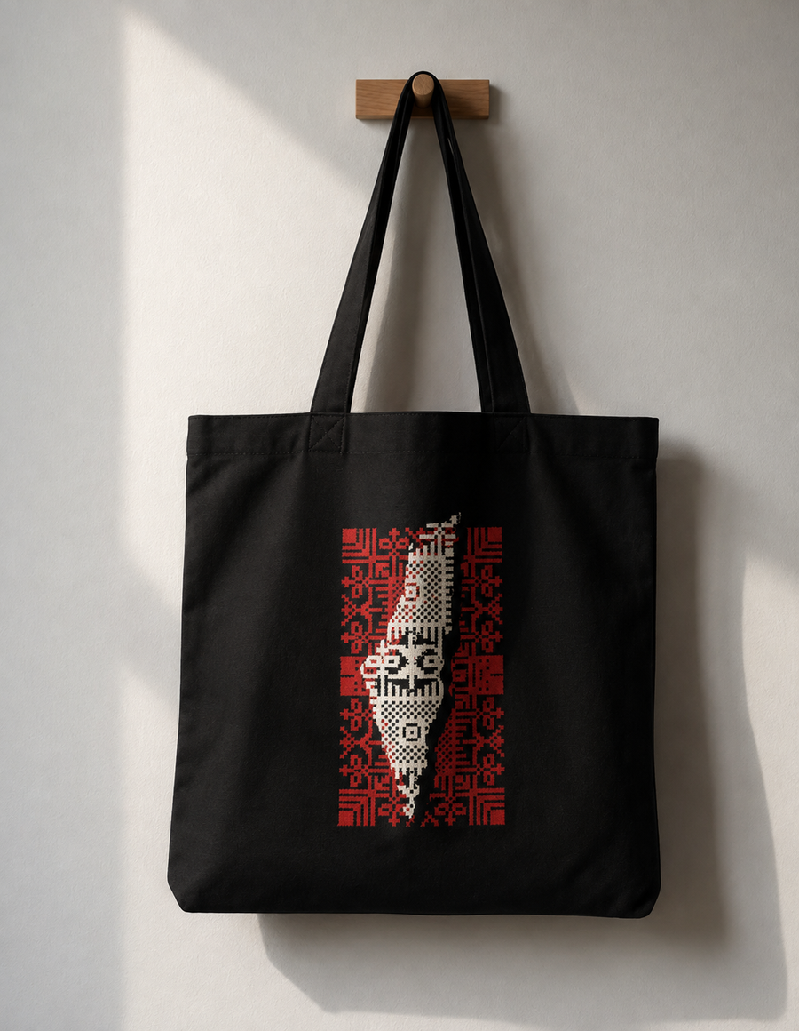 A black canvas tote bag hanging from a wooden peg on a light gray wall. The bag features a vertical graphic print of the Palestine map filled with a red and white pixelated pattern.