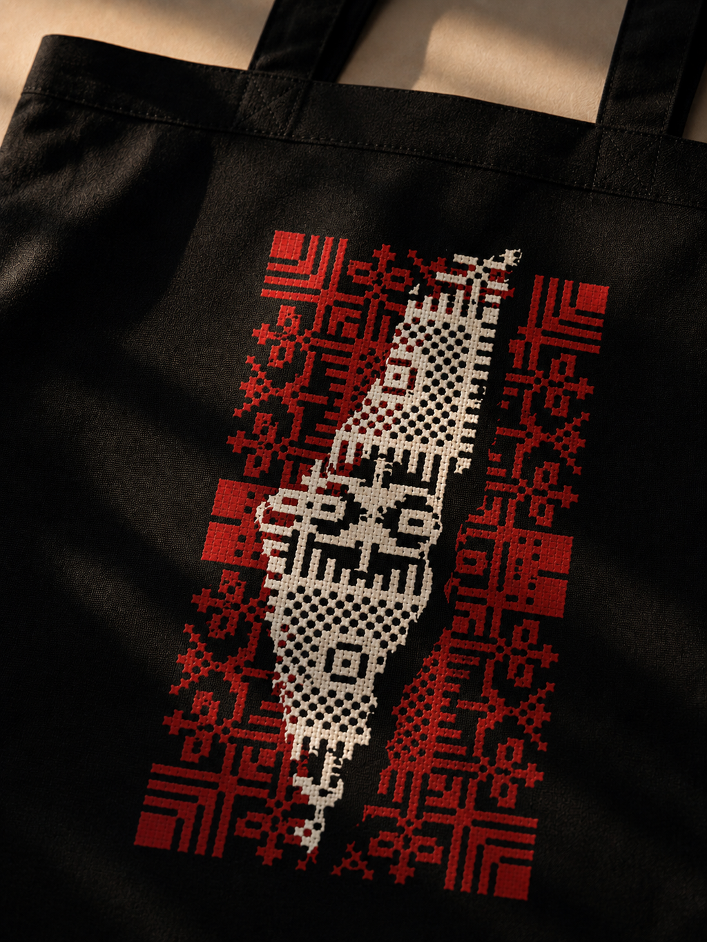 A close-up of a black tote bag featuring a stylized graphic of Palestine in white, flanked by red geometric patterns, set against a warm beige background.