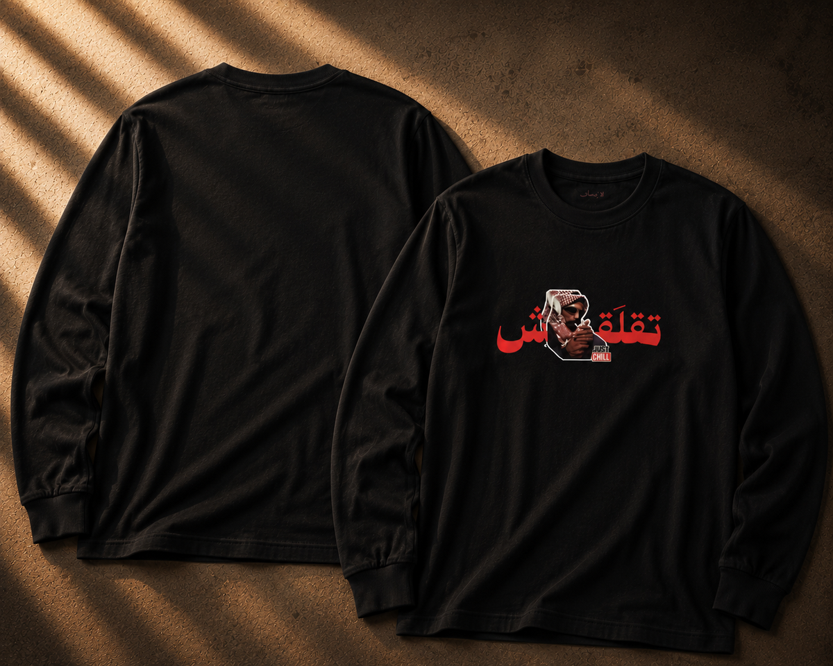 Two black long-sleeve t-shirts displayed on a textured brown surface with dramatic diagonal shadows. The left shirt shows the plain back, while the right shirt shows the front with a graphic print featuring red Arabic text and a small illustrated face.