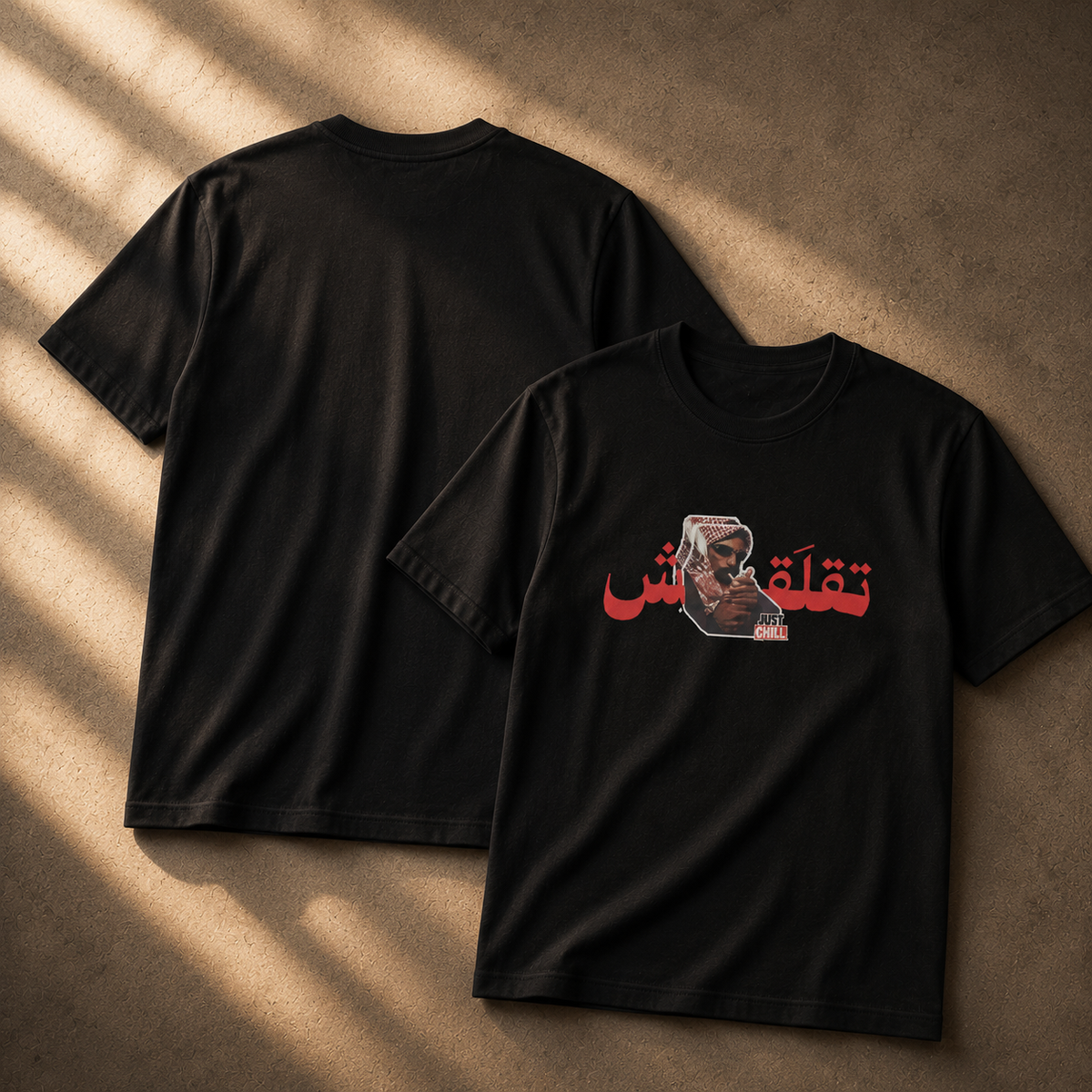 Two black short-sleeve T-shirts laid flat on a textured brown surface with diagonal warm light shadows. The front shirt shows a red Arabic script graphic and a small portrait print on the chest, while the back shirt is plain.