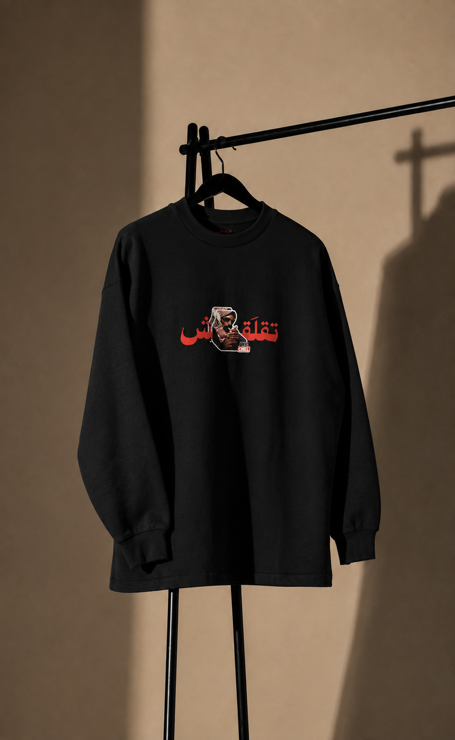 A black long sleeve tshirt hanging on a black clothing rack against a warm beige wall, featuring a graphic print on the chest with red Arabic text and a small portrait illustration.